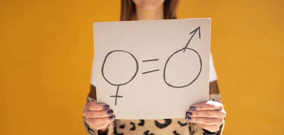 Frau steht vor gelbem Hintergrund. Sie hält ein Schild in die Kamera. Dort sind die beiden Geschlechtszeichen aufgemalt und durch zwei kurze parallele Striche getrennt.