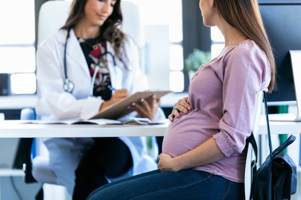 Eine schwangere Frau sitzt mit der Hand auf dem Bauch gegenüber einer Ärztin, die Notizen macht.  