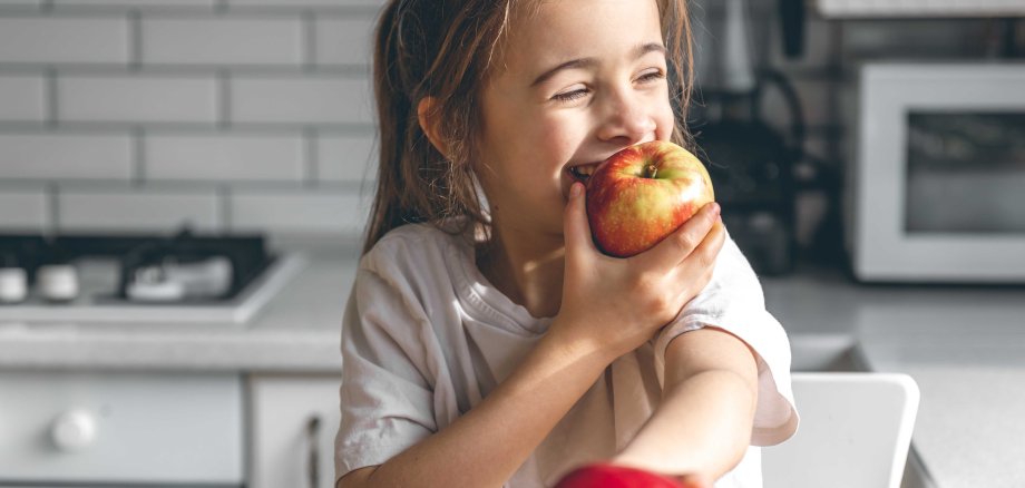 Kleines Mädchen beißt in einen Apfel. Im Hintergrund sieht man eine Küche.