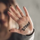 Im Hintergrund ein Portrait einer jungen Frau mit Blessuren im Gesicht. Im Vordergrund zeigt sie ihre Handinnenfläche, auf der in schwarzer Schrift "Stop!" steht. 