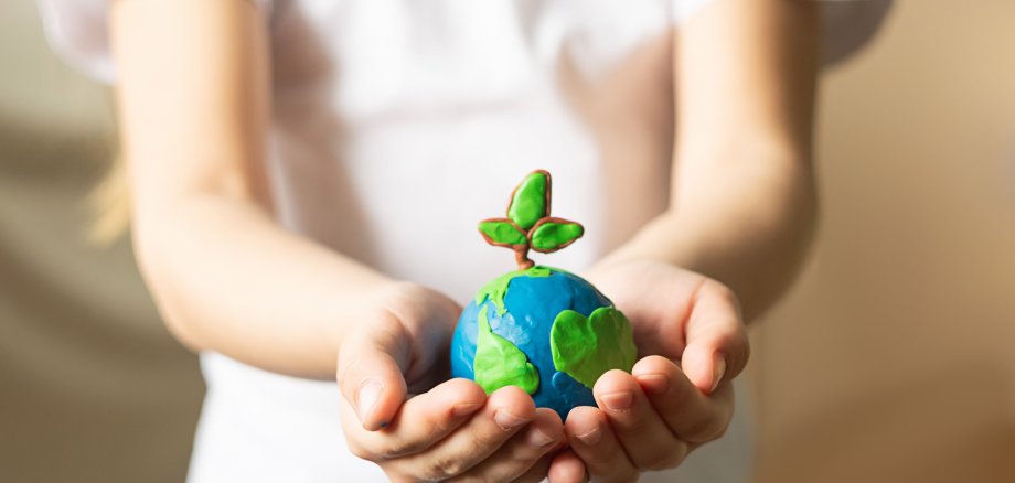 Ein kleiner blau-grüner Globus aus Wachs mit einem Wachspflanzenzwei darauf, in den Händen eines kleinen Mädchens.