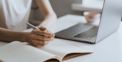 Eine Person sitzt an einem Tisch mit einem geöffneten Laptop und macht handschriftliche Notizen in einem Notizbuch. Im Hintergrund ist eine Tasse zu sehen.