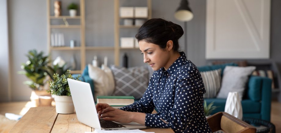 Frau im Seitenprofil sitzt auf einem Holzstuhl an einem Holztisch an ihrem silbernen Laptop und tippt. Neben ihr liegen Bücher und ein aufgeschlagenes Notizheft. Sie sitzt im Homeoffice in ihrem Wohnzimmer.