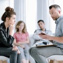Rebellierende Teenagerin hört einem Sozialpädagogen zu während einer Gruppentherapiestunde mit anderen Jugendlichen, die Probleme haben.
