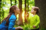 Ein Kind und eine Frau pusten gemeinsam eine Pusteblume im sonnigen Wald.