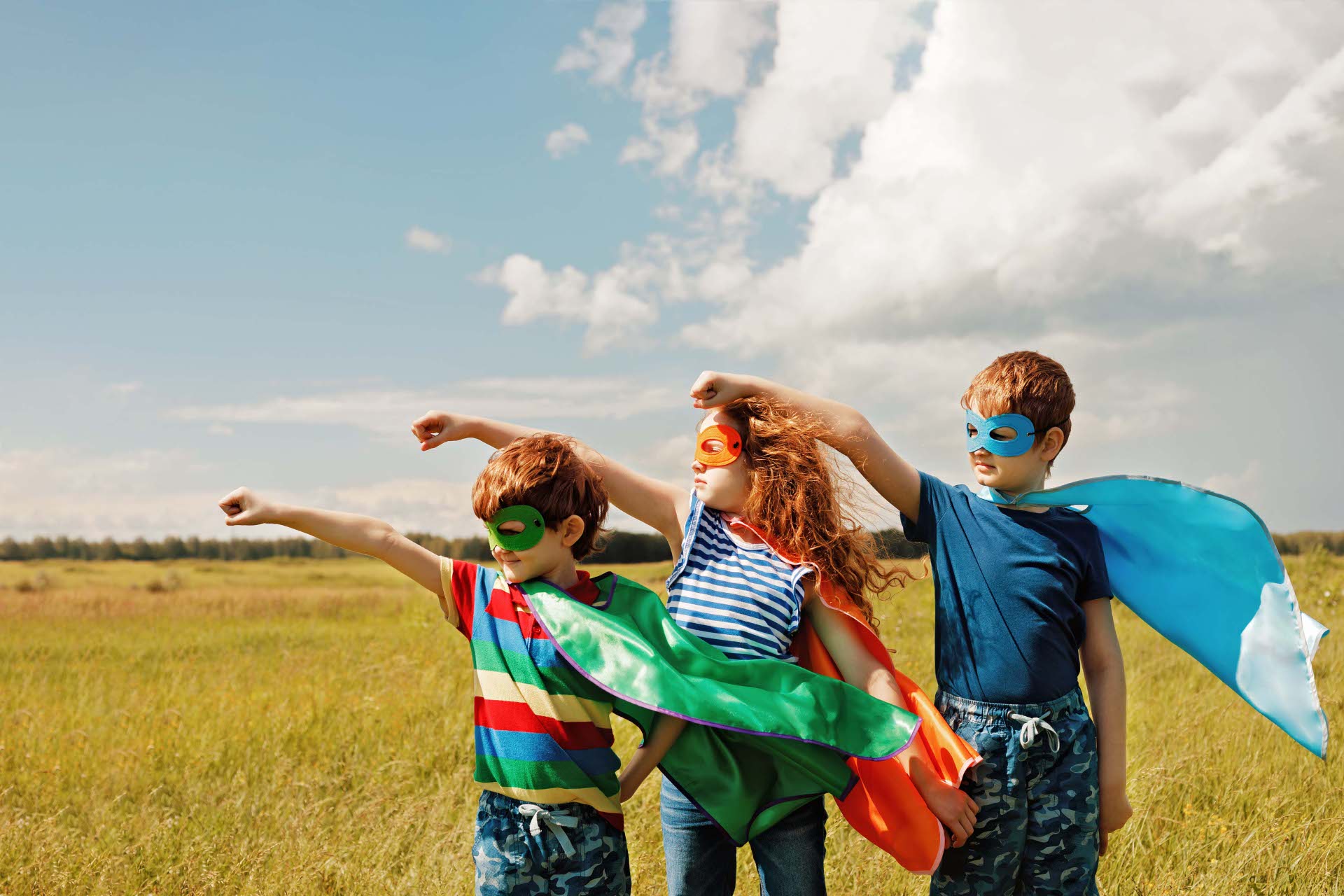 Drei Kinder in bunten Superheldenkostümen mit Umhängen und Masken posieren auf einer Wiese unter blauem Himmel und strecken die Fäuste in die Luft.