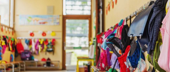 Bunte Kinderjacken und Rucksäcke hängen an Haken in einem Flur einer Kita oder Grundschule.