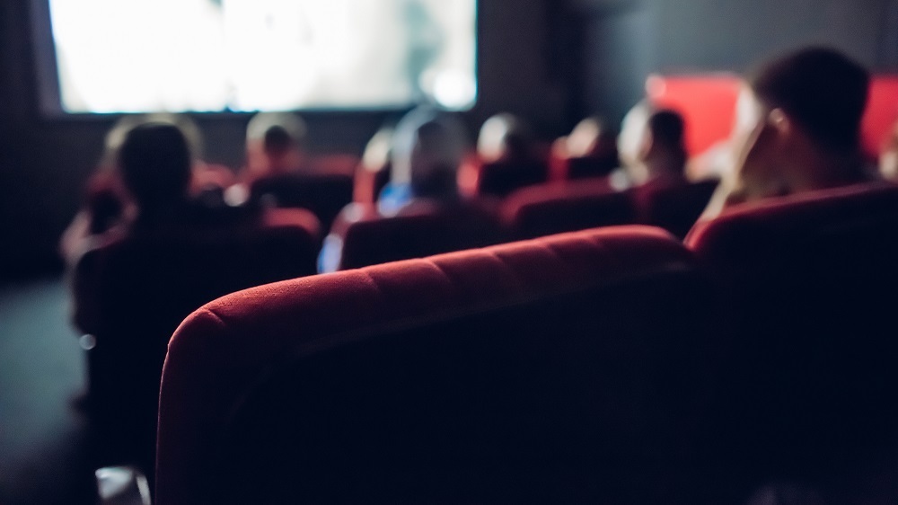 Rote Sitzplätze eines Kinos im Vordergrund. Im Hintergrund sieht man Leute und die Kino-Leinwand.