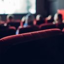 Rote Sitzplätze eines Kinos im Vordergrund. Im Hintergrund sieht man Leute und die Kino-Leinwand.