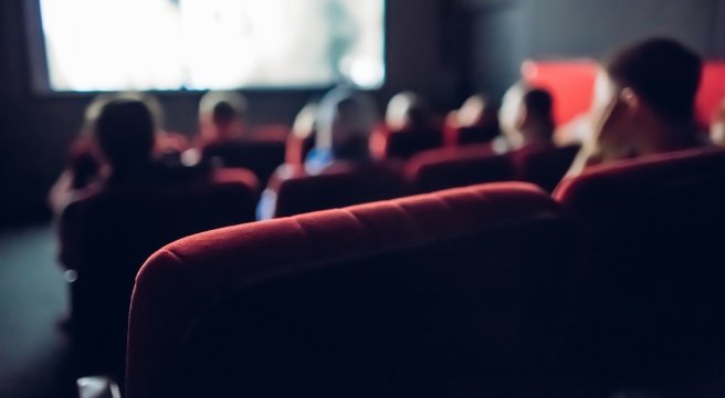 Rote Sitzplätze eines Kinos im Vordergrund. Im Hintergrund sieht man Leute und die Kino-Leinwand.