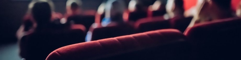 Rote Sitzplätze eines Kinos im Vordergrund. Im Hintergrund sieht man Leute und die Kino-Leinwand.