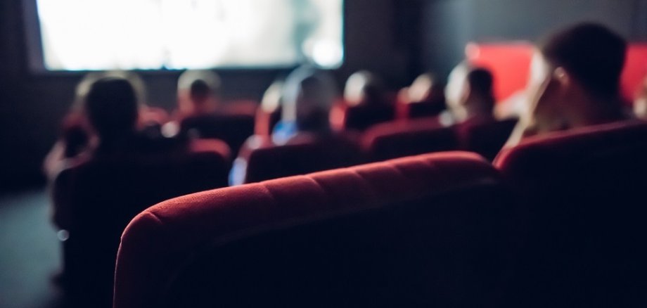 Rote Sitzplätze eines Kinos im Vordergrund. Im Hintergrund sieht man Leute und die Kino-Leinwand.