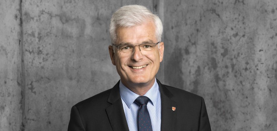 Porträt eines freundlich lächelnden Mannes mit kurzen weißen Haaren und Brille in dunklem Anzug, hellblauem Hemd und Krawatte vor grauem Hintergrund.