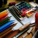 Auf einem Tisch liegen Buntstifte in vielen verschiedenen Farben und ein bunter Tuschkasten. Weitere Utensilien zum Malen, wie ein Pinsel und Farben stehen ebenfalls auf dem Tisch.