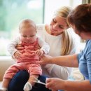  Das Bild zeigt zwei Frauen und ein Baby in einem Innenraum mit Tageslicht. Die Frau auf der linken Seite hält das Baby liebevoll im Arm, das eine rosafarbene Latzhose und ein weißes Shirt trägt. Die Frau auf der rechten Seite, die etwas größere Statur hat und ein blaues Oberteil trägt, scheint dem Baby etwas zu erklären oder zu zeigen, da sie dabei ist, mit ihrer Hand zu gestikulieren. Die Stimmung wirkt entspannt und fürsorglich.