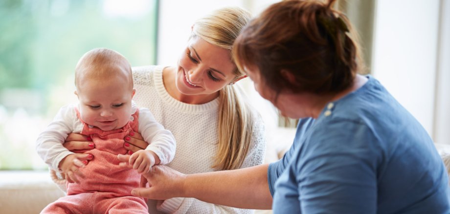  Das Bild zeigt zwei Frauen und ein Baby in einem Innenraum mit Tageslicht. Die Frau auf der linken Seite hält das Baby liebevoll im Arm, das eine rosafarbene Latzhose und ein weißes Shirt trägt. Die Frau auf der rechten Seite, die etwas größere Statur hat und ein blaues Oberteil trägt, scheint dem Baby etwas zu erklären oder zu zeigen, da sie dabei ist, mit ihrer Hand zu gestikulieren. Die Stimmung wirkt entspannt und fürsorglich.