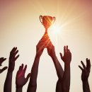 Viele Hände halten gemeinsam einen Pokal in die Höhe. Im Hintergrund geht die Sonne unter.
