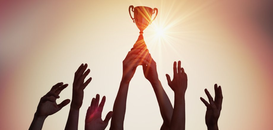 Viele Hände halten gemeinsam einen Pokal in die Höhe. Im Hintergrund geht die Sonne unter.