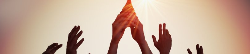 Viele Hände halten gemeinsam einen Pokal in die Höhe. Im Hintergrund geht die Sonne unter.