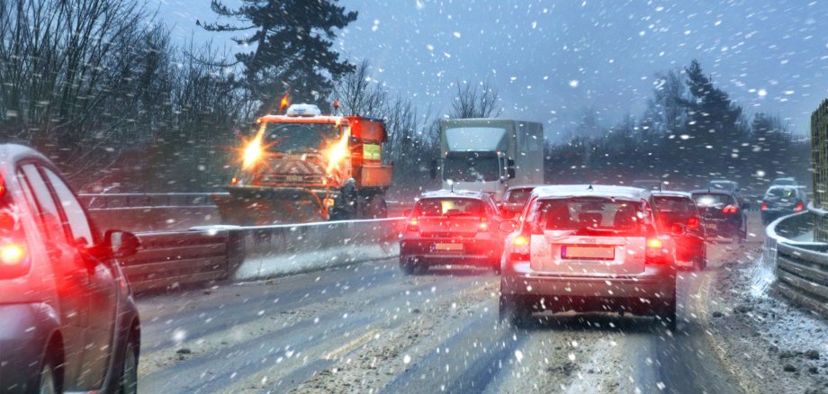 Einige Fahrzeuge fahren auf einer schneebedeckten Autobahn. Es schneit. Auf der linken Fahrspur ist zudem ein Streu- und Räumfahrzeug zu sehen.