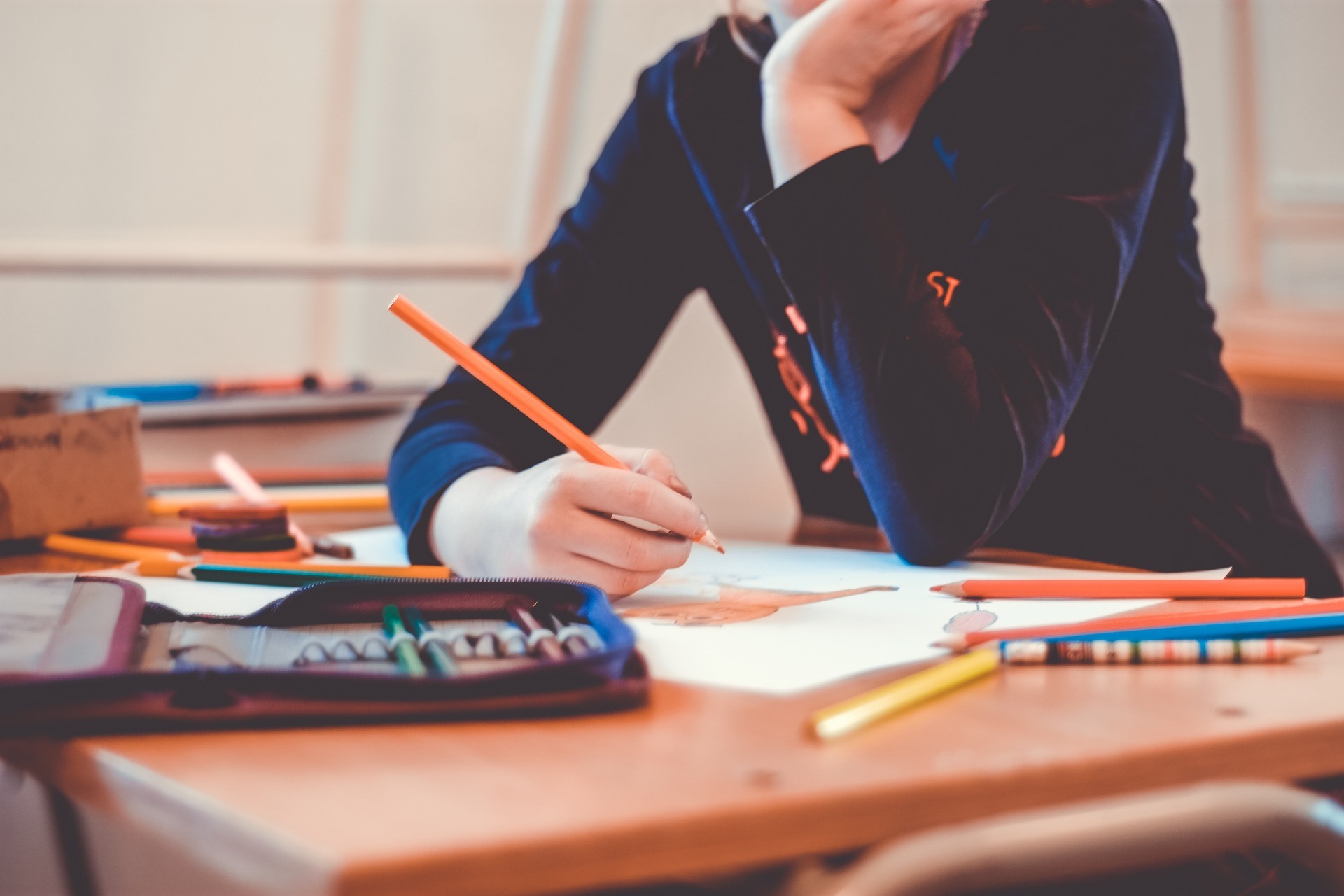 Nahaufnahme einer Schulbank, an der eine Schülerin sitzt und mit Buntstiften ein Bild malt. Auf der Schulbank liegen eine Federmappe und viele Buntstifte.