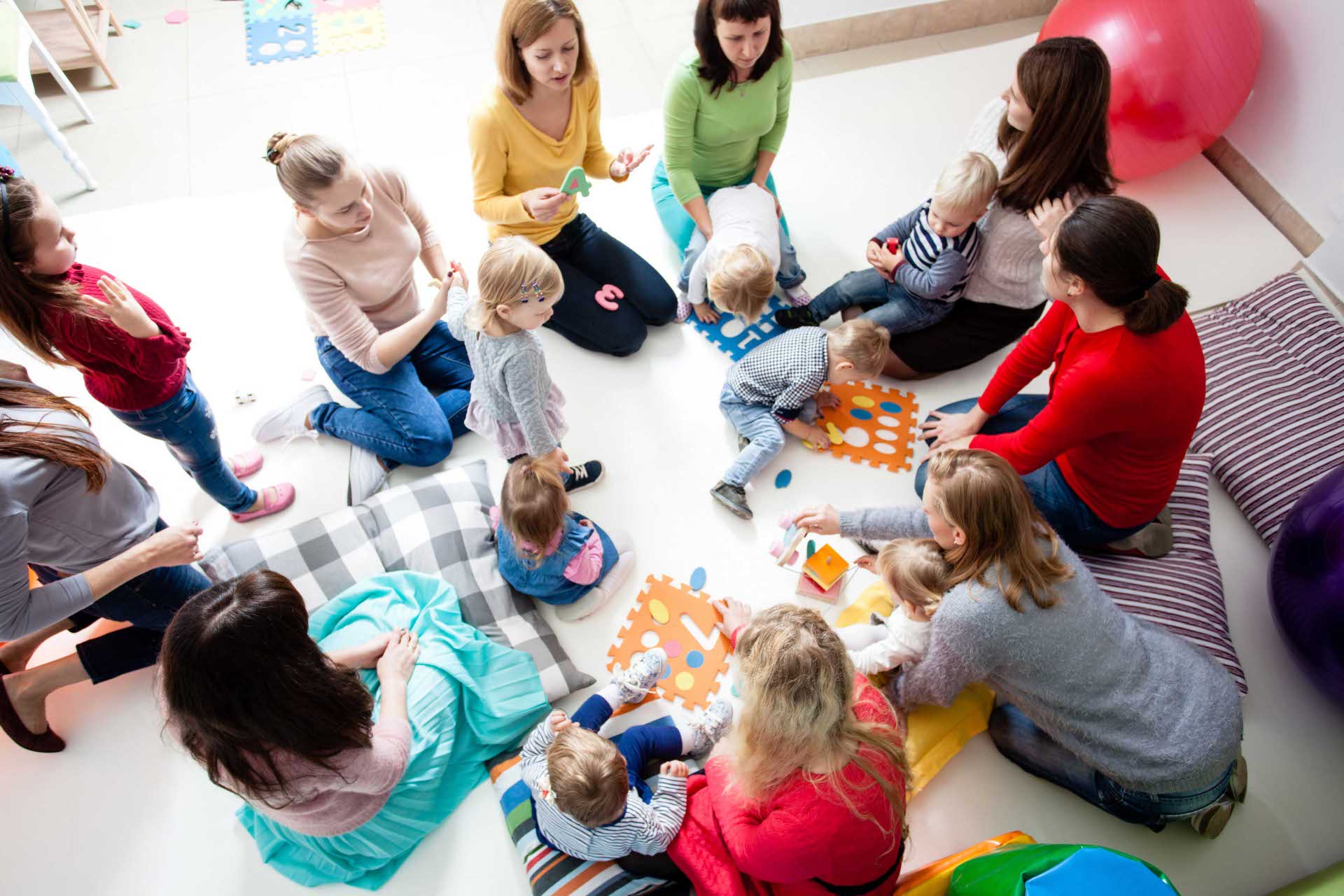 Mehrere Eltern und Kleinkinder sitzen auf dem Boden und spielen mit bunten Spielmaterialien.