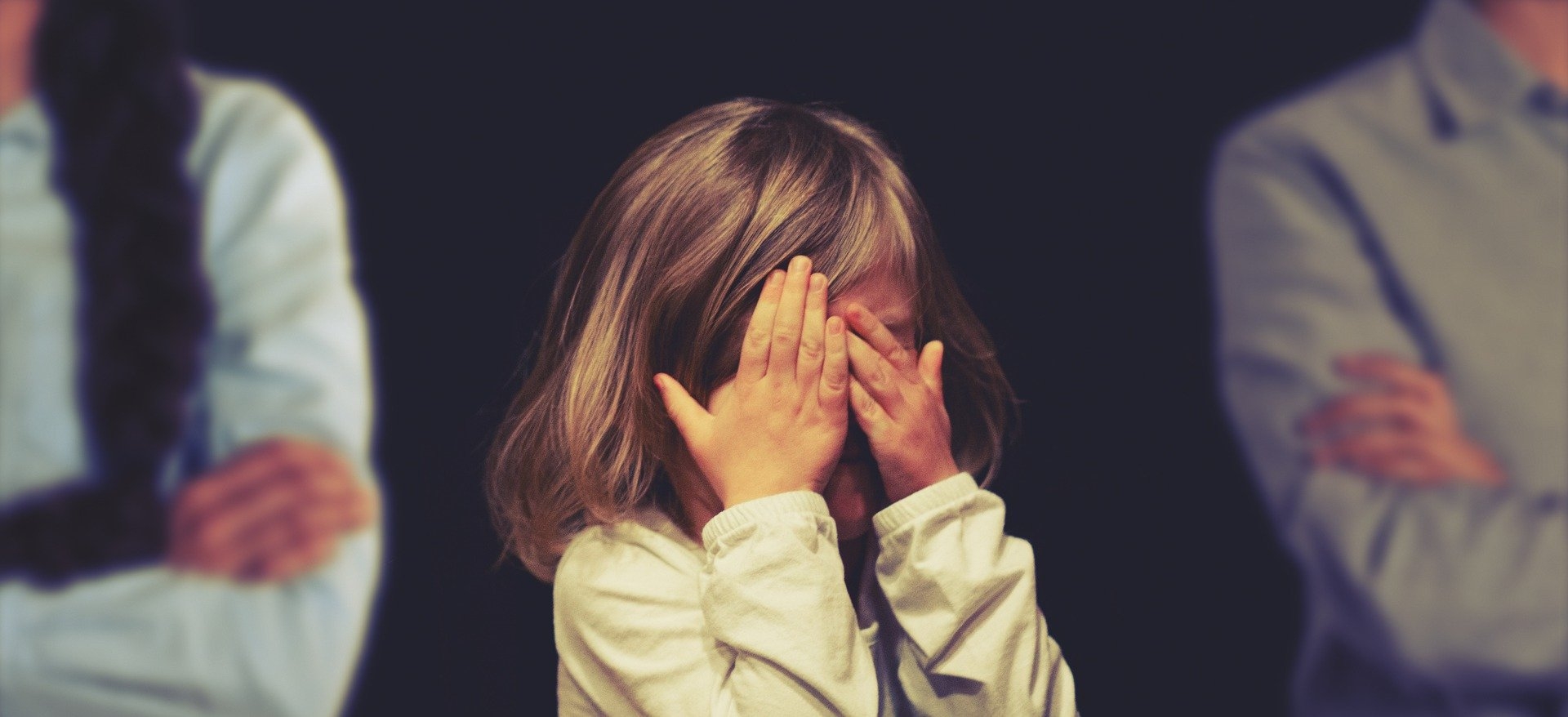 Kleines trauriges Mädchen hält sich die Hände vor das Gesicht. Im schwarzen Hintergrund sind rechts und links jeweils der Oberkörper einer Frau und eines Mannes zur Hälfte zu sehen.