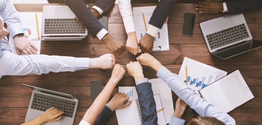 Mehrere Angestellte sitzen an einem Büro-Tisch. Sie haben Laptops und Blätter vor sich liegen und strecken ihre Faust zum Gruß in die Mitte. Das Foto ist von oben aufgenommen.