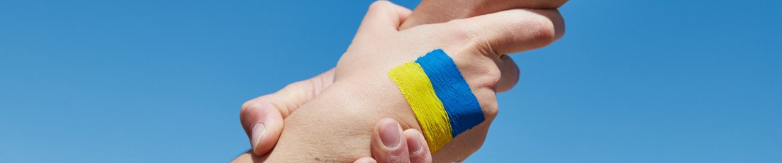 Eine Hand hält eine andere Hand fest- Auf dem Handrücken gemalt ist die ukrainische Flagge in blau-gelb gemalt. Blauer Hintergrund.