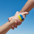 Eine Hand hält eine andere Hand fest- Auf dem Handrücken gemalt ist die ukrainische Flagge in blau-gelb gemalt. Blauer Hintergrund.