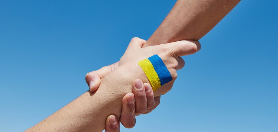 Eine Hand hält eine andere Hand fest- Auf dem Handrücken gemalt ist die ukrainische Flagge in blau-gelb gemalt. Blauer Hintergrund.
