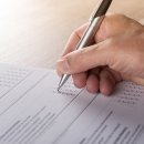 Eine männliche Hand hält einen Stift. Auf dem Tisch liegt ein Blatt Papier. Auf dem Blatt wird mit dem Stift ein Haken auf einen Kasten gesetzt.
