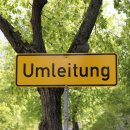Ein gelbes Schild mit der schwarzen Aufschrift "Umleitung" steht vor einem Baum. Im Hintergrund sind viele grüne Blätter von weiteren Bäumen sichtbar.
