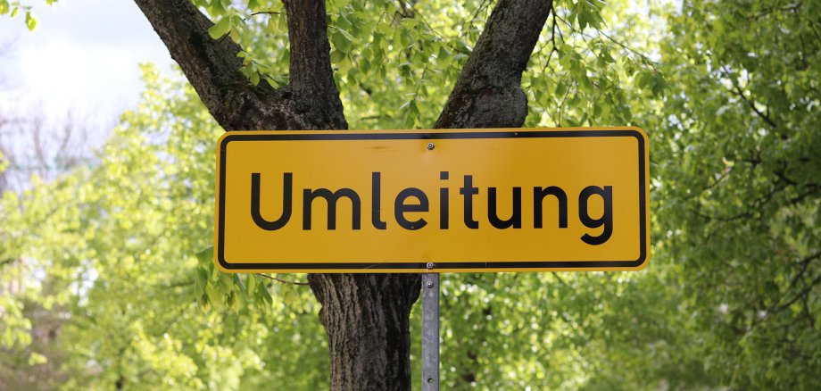 Ein gelbes Schild mit der schwarzen Aufschrift "Umleitung" steht vor einem Baum. Im Hintergrund sind viele grüne Blätter von weiteren Bäumen sichtbar.