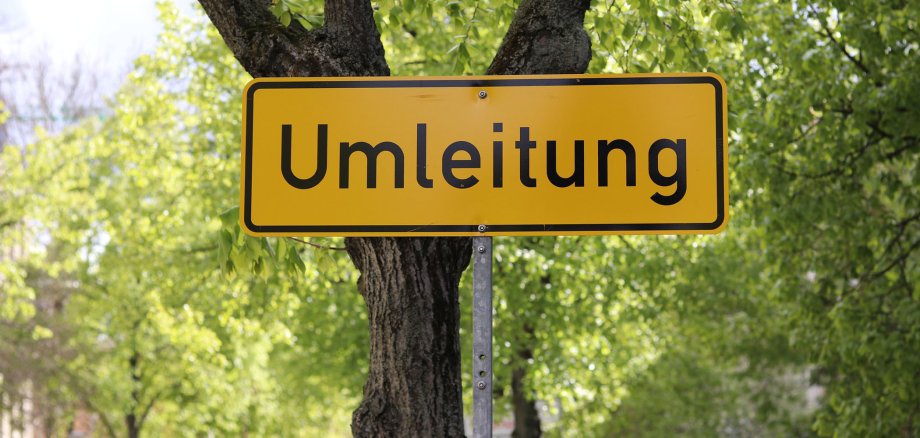 Ein gelbes Schild mit der schwarzen Aufschrift "Umleitung" steht vor einem Baum. Im Hintergrund sind viele grüne Blätter von weiteren Bäumen sichtbar.