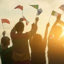 Einige Menschen schwenken Fahnen verschiedener Länder in der Luft. Es ist Sonnenuntergang, die Menschen stehen mit dem Rücken zur Kamera. 