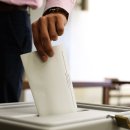 Eine männliche Hand wirft einen Wahlzettel in eine Wahlurne. Im Hintergrund ist eine Wahlkabine zu sehen.