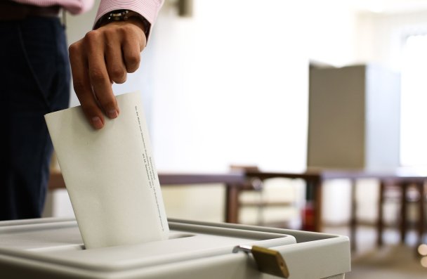 Eine männliche Hand wirft einen Wahlzettel in eine Wahlurne. Im Hintergrund ist eine Wahlkabine zu sehen.