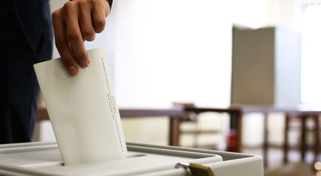 Eine männliche Hand wirft einen Wahlzettel in eine Wahlurne. Im Hintergrund ist eine Wahlkabine zu sehen.