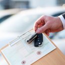 Die Hand eines Autoverkäufers halt einen Schlüssel und die Zulassungs-Papiere in der Hand. Im Hintergrund stehen Autos.