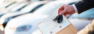 Die Hand eines Autoverkäufers halt einen Schlüssel und die Zulassungs-Papiere in der Hand. Im Hintergrund stehen Autos.