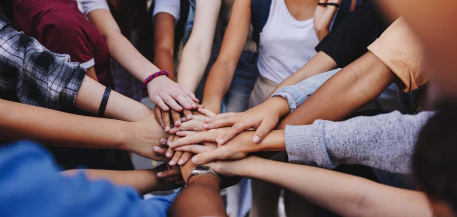 Mehrere Jugendliche legen im Kreis stehend ihre Hände übereinander in die Mitte – ein Zeichen für Zusammenhalt, Teamgeist und gemeinsames Engagement. Die Szene zeigt Vielfalt und Verbundenheit.