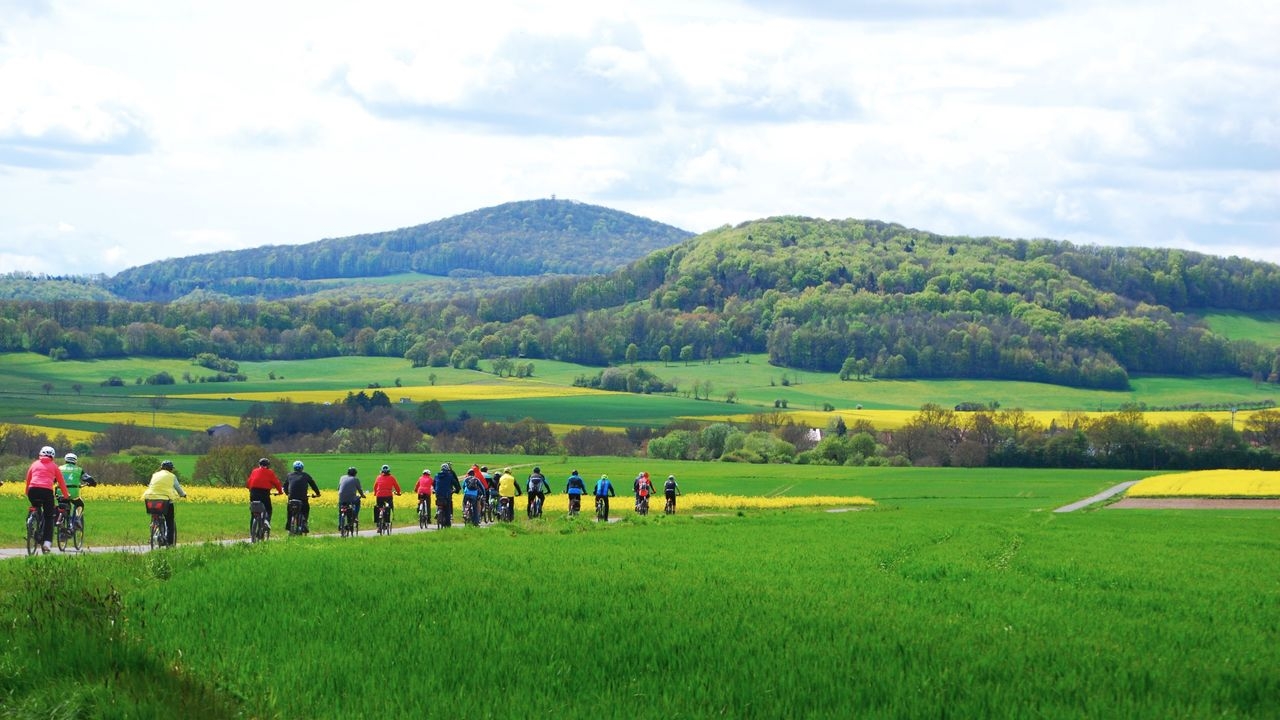 Eine Gruppe von Radfahrenden fährt in bunten Jacken auf einem Feldweg durch die Kuppenrhön, umgeben von grünen Wiesen, blühenden Rapsfeldern und bewaldeten Hügeln.