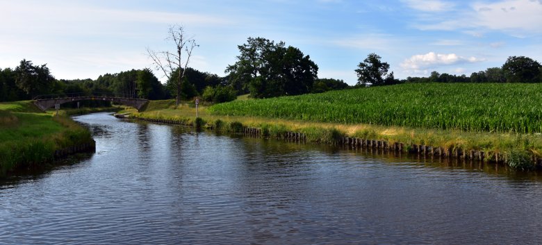 Ein Fluss, der sich durch eine grüne Landschaft zieht. Im Hintergrund ist eine Steinbrücke über dem Fluss zu sehen, weiter hinten grüne Bäume. Blauer Himmel mit Schleierwolken.