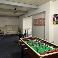In einem Raum steht ein Sofa aus Paletten. An der Wand hängt ein Fernseher. In einer Ecke steht eine Musikanlage. An einer anderen Wand hängt eine Dartscheibe.
