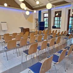 In einem großen, hellen Saal stehen ungefähr 30 Stühle in fünf Reihen. Vor den Stühlen stehen zwei Flipcharts, ein Beamer und ein Rednertisch.