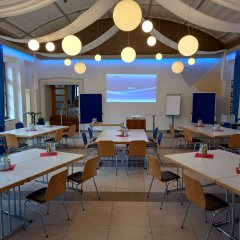 In einem hellen großen Saal stehen fünf Tische mit jeweils sechs Stühlen. Auf den Tischen stehen Getränke. Vor den Stühlen stehen ein Flipchart, ein Beamer und zwei Pinnwände. Auf der Wand wird das Bild des Beamer angestrahlt.