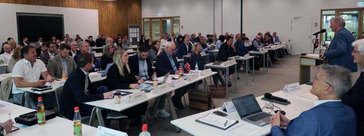 Ein großer Konferenzraum mit zahlreichen Teilnehmenden an U-förmig angeordneten Tischen. Im Vordergrund sitzt das Präsidium mit Laptop und Unterlagen, während rechts eine Person an einem Rednerpult einen Vortrag hält. Die Zuhörenden verfolgen die Präsentation aufmerksam