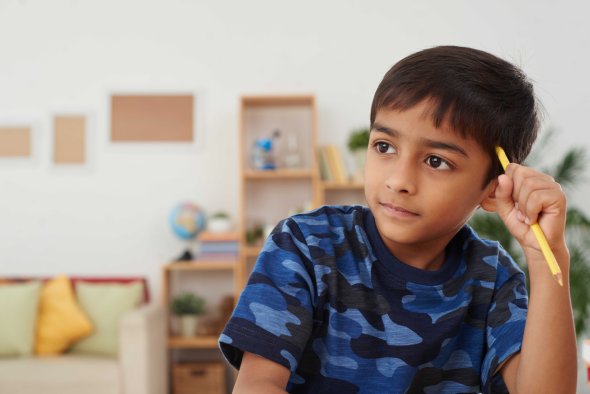 Ein nachdenklicher Junge in einem blauen Tarnmuster-T-Shirt hält einen Bleistift an den Kopf, während er in einem Raum mit Bücherregalen im Hintergrund sitzt.