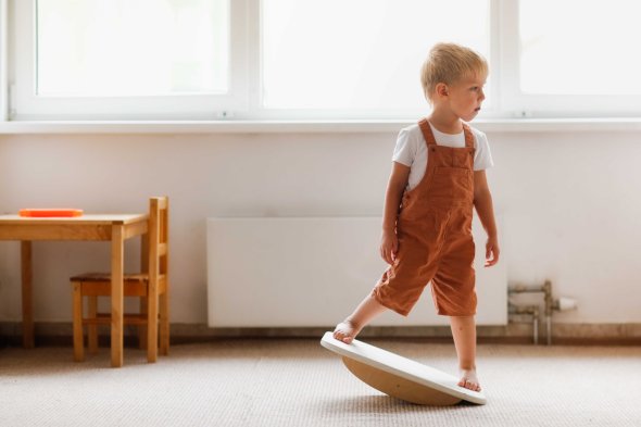 Ein kleiner Junge in brauner Latzhose steht barfuß auf einem Balancebrett in einem hellen Raum mit Holzmöbeln.